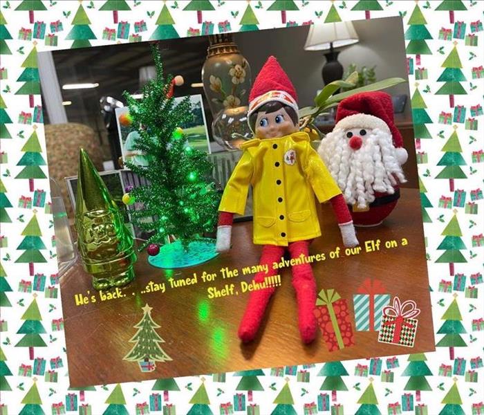 Photo shows elf on the shelf in a yellow raincoat with text overlay stating that he is back and to stay tuned.